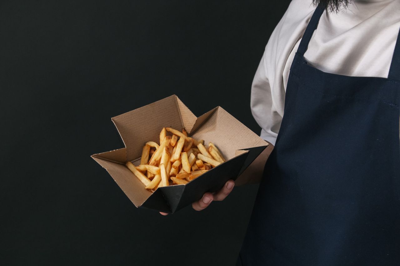 An open cardboard food carton holding french fries, held by a person in an apron