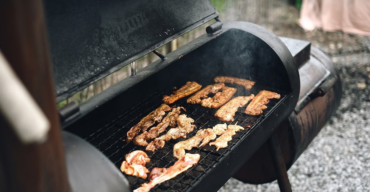 Barrel smoker with the lid open, strips of meat on the grate and smoke rising from the chamber