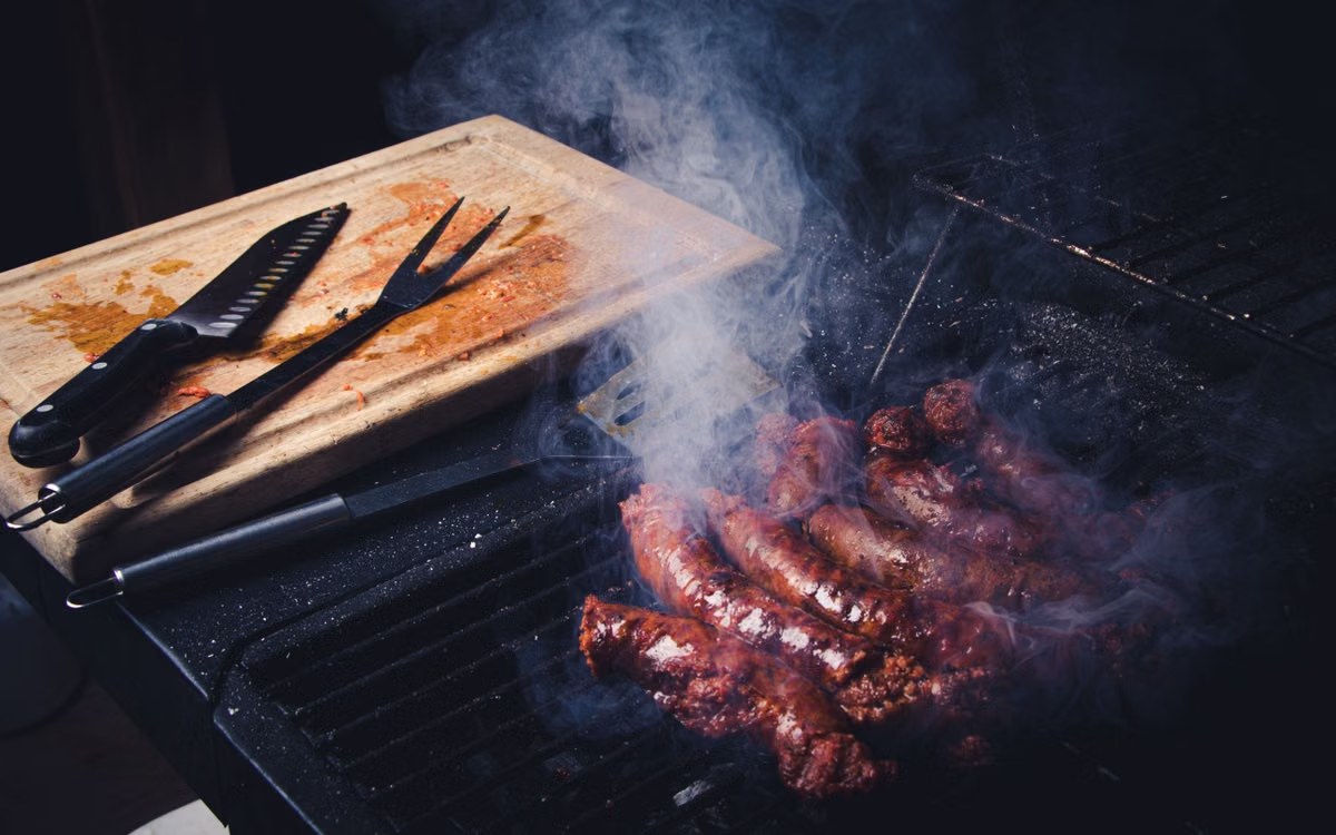 Sausages smoking on a barbecue grate with a wooden board and carving tools alongside