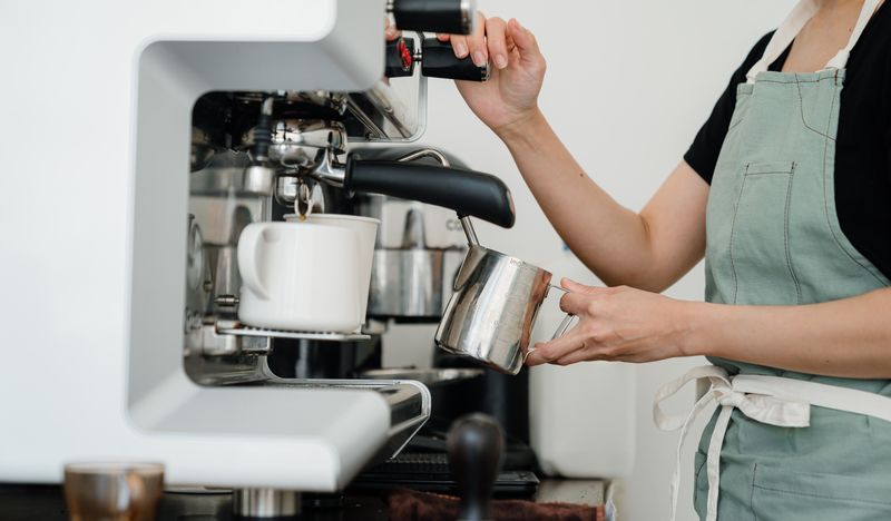 A barista steaming milk in a jug with the steam wand of a white espresso machine