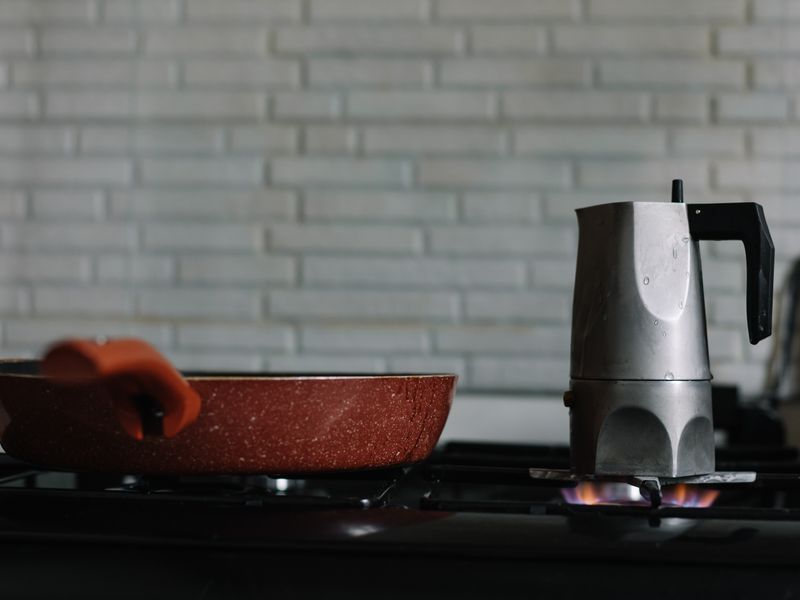 Moka pot on a flat black stovetop beside a red frying pan